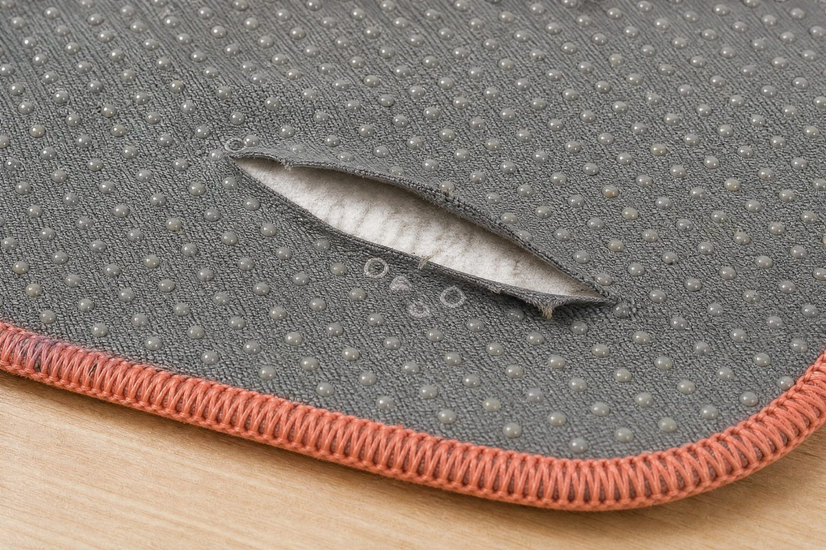 Close-up of the grey silicone-dot underside of a worn washable pee pad, one finger-length split lifting to show the white core, the overlocked edge beside it