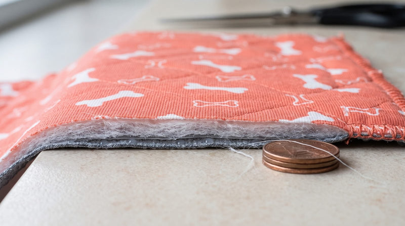 A washable pee pad cut across to show its four layers: the printed face, the white absorbent core, the thin waterproof film and the grey silicone-dot underside, with two coins beside the cut edge for thickness.